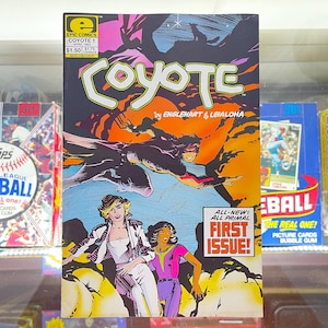 May include: A vintage comic book titled "Coyote" with a vibrant cover illustration. The cover features a dynamic action scene with figures in a desert landscape. The comic is labeled "First Issue" and is surrounded by baseball and football card boxes.