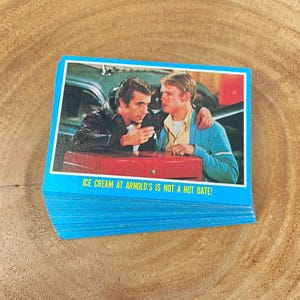 May include: A stack of blue trading cards featuring a scene with two men and the text "ICE CREAM AT ARNOLD'S IS NOT A HOT DATE!". The image shows two men in a diner setting.