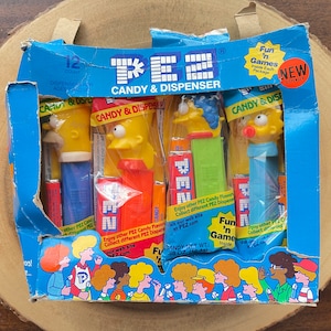 May include: A box of 12 Simpsons PEZ dispensers. The box is blue and features a picture of the Simpsons characters. The dispensers are individually packaged and include a variety of characters, including Homer, Marge, Bart, and Lisa. The box also includes a small sticker that says "Fun 'n Games Inside".