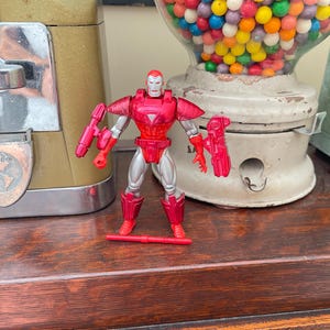 Pode incluir: Uma figura de a&ccedil;&atilde;o do Homem de Ferro vermelha e prateada com um acess&oacute;rio de arma vermelho. A figura est&aacute; em uma superf&iacute;cie de madeira, com uma m&aacute;quina de goma de mascar vintage e outros objetos ao fundo.