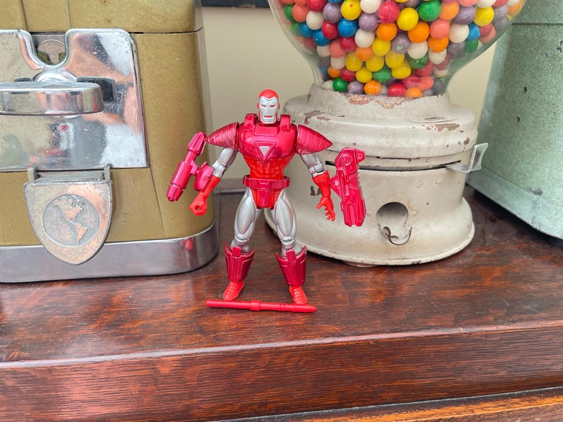Pode incluir: Uma figura de a&ccedil;&atilde;o do Homem de Ferro vermelha e prateada com um acess&oacute;rio de arma vermelho. A figura est&aacute; em uma superf&iacute;cie de madeira, com uma m&aacute;quina de goma de mascar vintage e outros objetos ao fundo.