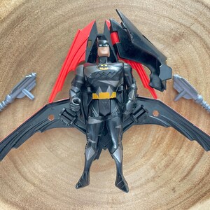 May include: A Batman action figure with a black and gray suit, yellow belt, and a black and red wing apparatus. Two gray toy guns are positioned on either side of the figure. The figure is on a wooden surface.