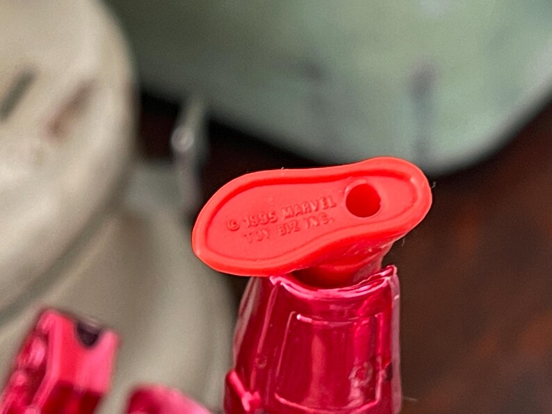 Pode incluir: Close-up de um p&eacute; de brinquedo de pl&aacute;stico vermelho com um buraco. O texto "&copy; 1995 MARVEL" &eacute; vis&iacute;vel. O brinquedo faz parte de uma figura maior, com um torso vermelho e outras partes vis&iacute;veis no fundo.