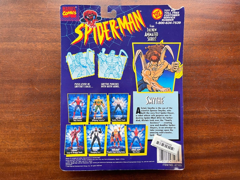 May include: A blue and purple action figure card featuring Spider-Man and the villain Smythe. The card has a picture of Smythe with his arms outstretched and a description of his character. The card also shows different action poses for Spider-Man and Smythe.