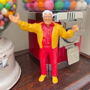 May include: A vintage figurine of a man in a red shirt, red pants, and yellow jacket, with arms outstretched. The figurine is positioned in front of a red gumball machine with a silver coin slot. A vintage gumball machine is on the left.