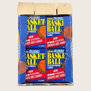 May include: A cardboard display box containing four individual packs of 1991 Fleer basketball player photo cards. Each pack features blue, red, and yellow text with a basketball graphic, labeled with New Pro Visions Sports Art Cards and 14 cards.