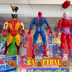 May include: Three action figures of Marvel comic book characters: Wolverine, Spider-Man, and Magneto. Wolverine is yellow and red, Spider-Man is blue and red, and Magneto is red and purple. The figures are displayed in front of a baseball and basketball card display.