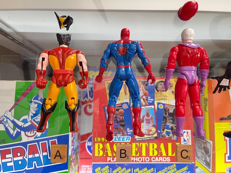 May include: Three action figures of Marvel comic book characters: Wolverine, Spider-Man, and Magneto. Wolverine is yellow and red, Spider-Man is blue and red, and Magneto is red and purple. The figures are displayed in front of a baseball and basketball card display.
