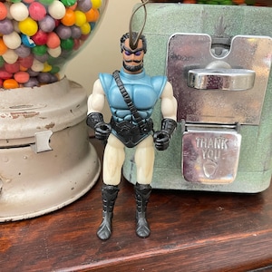 May include: A blue and white action figure with a black belt and boots. The figure has a black mask and a black antenna on its head. The figure is standing on a wooden surface.