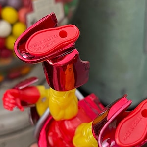 Może przedstawiać: Zbliżenie czerwono-ż&oacute;łtej figurki z metalicznymi akcentami. Figurka ma napis "&copy;1995 MARVEL TOY BIZ INC." na stopie. Figurka znajduje się przed automatem do gumy do żucia.