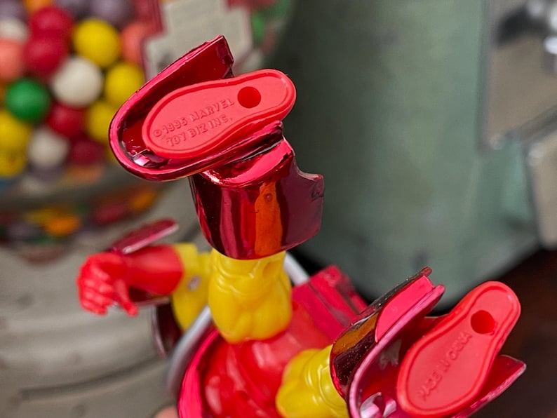 Może przedstawiać: Zbliżenie czerwono-ż&oacute;łtej figurki z metalicznymi akcentami. Figurka ma napis "&copy;1995 MARVEL TOY BIZ INC." na stopie. Figurka znajduje się przed automatem do gumy do żucia.