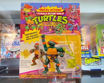 May include: A Teenage Mutant Ninja Turtles wind-up toy featuring Michelangelo, the green turtle with an orange bandana. The toy has a motorized rotating wrist action and comes with a sewer cover, nunchucks, and sewer snakes. The packaging features the Teenage Mutant Ninja Turtles logo and the words "Wacky Action".