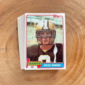 May include: A stack of Topps trading cards featuring Archie Manning, quarterback for the New Orleans Saints. The card shows Manning in his gold helmet and black and gold uniform. The card is on a wooden surface.