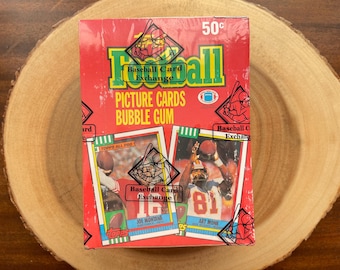 May include: A red, green, and white box of Topps Football Picture Cards and Bubble Gum. The box features a picture of Joe Montana and Art Monk, both wearing their football uniforms. The box also includes the text "Baseball Card Exchange" and "50c".