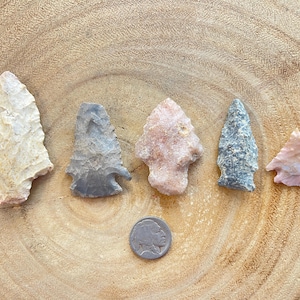 May include: Five arrowheads of different colors and shapes, including gray, white, pink, and brown, are arranged in a row on a wooden surface. A silver coin is in the center of the image.