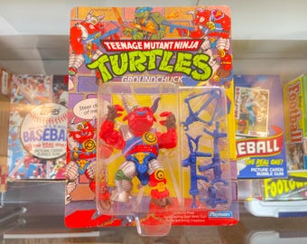 May include: Teenage Mutant Ninja Turtles action figure of Groundchuck, a red and silver bull with a blue and yellow chest plate. The figure comes with a blue crossbow and other accessories. The figure is packaged in a plastic blister pack with a cardboard backing.