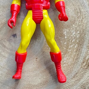 May include: A plastic Iron Man action figure, featuring a red and yellow color scheme. The figure's legs and arms are yellow, while the chest, boots, and gloves are red. The figure is posed on a wooden surface.