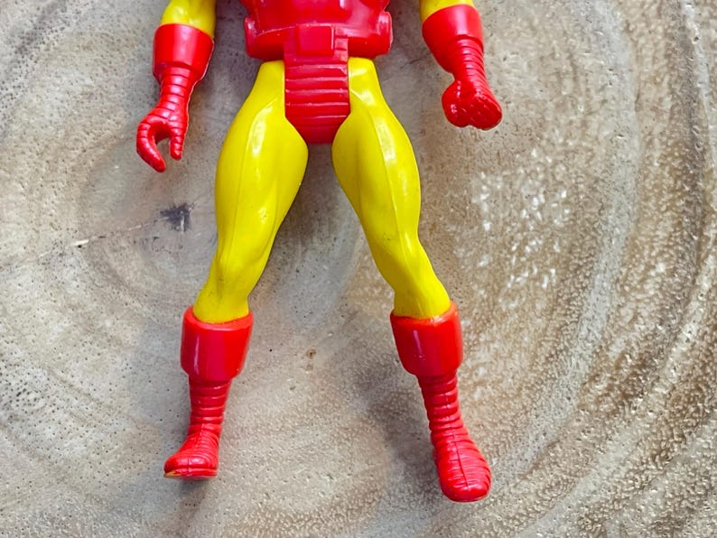 May include: A plastic Iron Man action figure, featuring a red and yellow color scheme. The figure's legs and arms are yellow, while the chest, boots, and gloves are red. The figure is posed on a wooden surface.