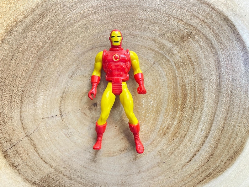 May include: A vintage Iron Man action figure. The figure is primarily yellow with red accents on the chest, boots, gloves, and helmet. The figure is standing on a wooden surface.