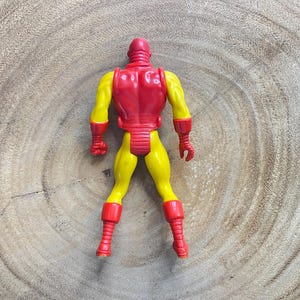May include: A vintage Iron Man action figure, viewed from the back, with a red helmet and armor, and yellow arms and legs. The figure has red gloves and boots. The toy is posed on a wooden surface.
