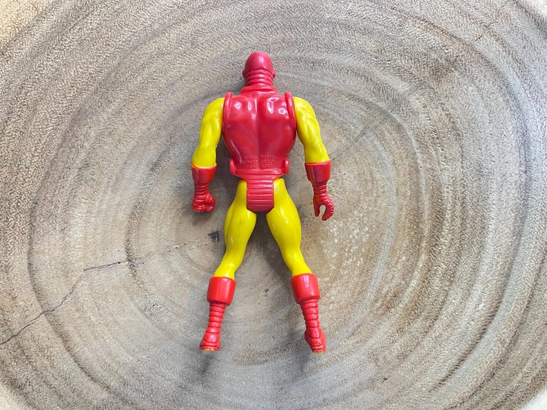 May include: A vintage Iron Man action figure, viewed from the back, with a red helmet and armor, and yellow arms and legs. The figure has red gloves and boots. The toy is posed on a wooden surface.
