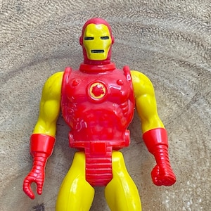 May include: A vintage Iron Man action figure. The toy features a red and yellow color scheme, with a yellow head and arms, and a red torso, gloves, and boots. The figure is posed upright, showcasing its classic design.