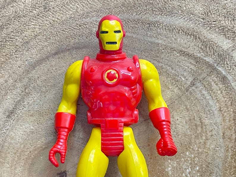 May include: A vintage Iron Man action figure. The toy features a red and yellow color scheme, with a yellow head and arms, and a red torso, gloves, and boots. The figure is posed upright, showcasing its classic design.