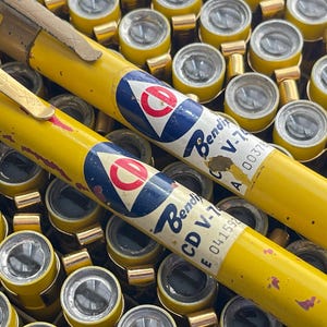 May include: Close-up of vintage yellow cylindrical objects with gold-colored metal ends. The objects have a blue and white label with a red circle and the text "Bendix V-7". The background is filled with similar objects.