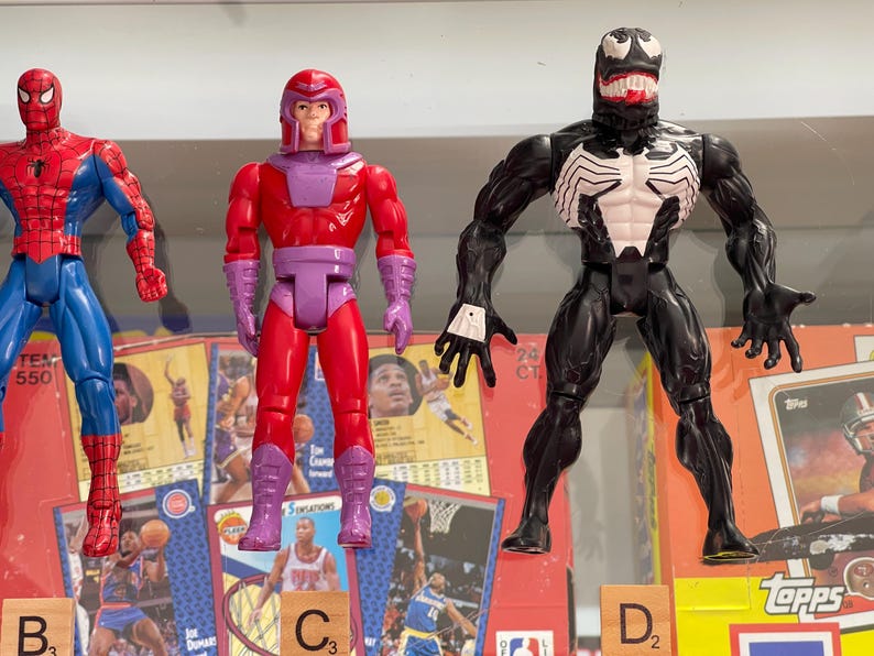 May include: Three action figures of Marvel comic book characters: Spider-Man in red and blue, Magneto in red and purple, and Venom in black and white. The figures are displayed in front of sports trading cards and wooden letter tiles.