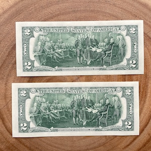 May include: Two $2 bills with a green background featuring a depiction of the signing of the Declaration of Independence. The text "THE UNITED STATES OF AMERICA" is printed at the top of each bill. The text "TWO DOLLARS" is printed at the bottom of each bill.
