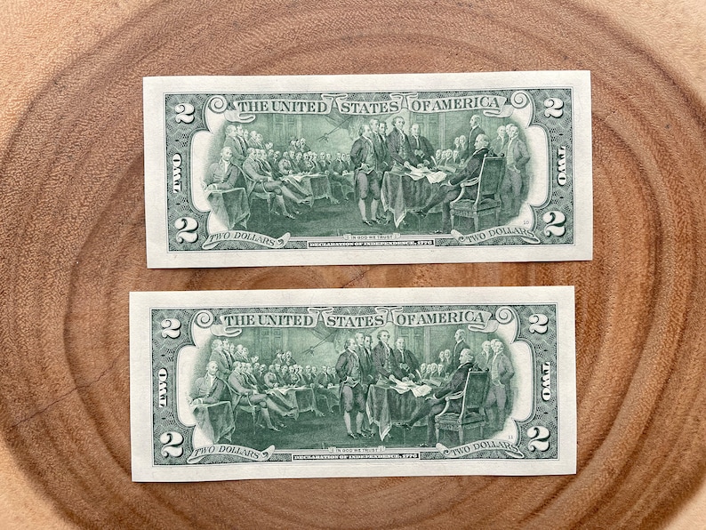 May include: Two $2 bills with a green background featuring a depiction of the signing of the Declaration of Independence. The text "THE UNITED STATES OF AMERICA" is printed at the top of each bill. The text "TWO DOLLARS" is printed at the bottom of each bill.