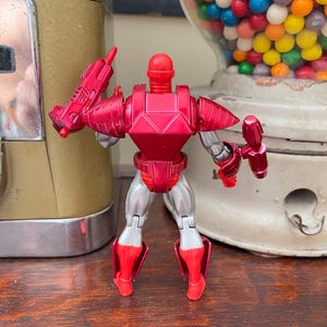 Pode incluir: Figura de a&ccedil;&atilde;o vermelha e prateada de um rob&ocirc; com capacete, armadura de peito e armadura de pernas. A figura est&aacute; segurando duas armas vermelhas. O rob&ocirc; est&aacute; em frente a uma m&aacute;quina de chicletes vintage e um dispensador de chicletes.