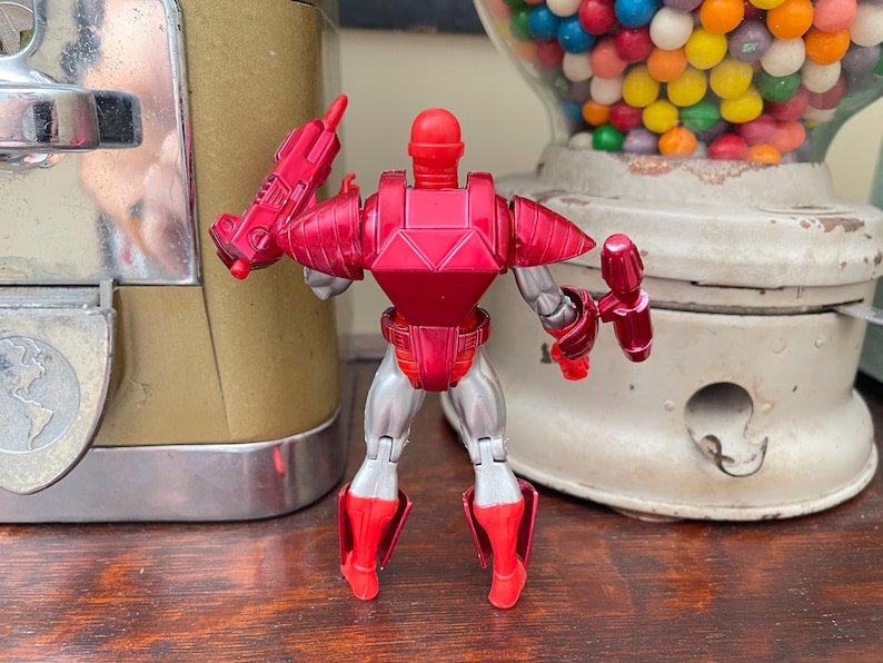 Pode incluir: Figura de a&ccedil;&atilde;o vermelha e prateada de um rob&ocirc; com capacete, armadura de peito e armadura de pernas. A figura est&aacute; segurando duas armas vermelhas. O rob&ocirc; est&aacute; em frente a uma m&aacute;quina de chicletes vintage e um dispensador de chicletes.