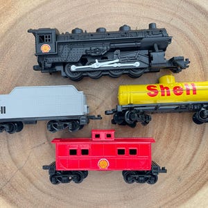 May include: A collection of toy train cars, including a black locomotive, a gray Shell car, a yellow Shell tanker car, and a red caboose. The Shell logo is visible on several cars. The toys are arranged on a wooden surface.