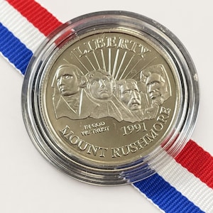 May include: A silver-toned commemorative coin featuring the Mount Rushmore monument with the text LIBERTY, IN GOD WE TRUST, 1991, and MOUNT RUSHMORE, encased in a clear protective plastic capsule and resting on a red, white, and blue striped ribbon.