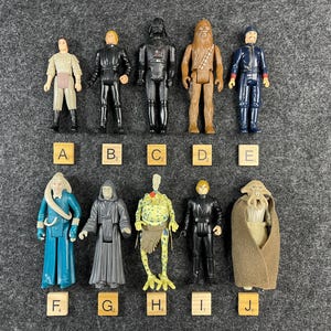 May include: A collection of ten vintage plastic action figures arranged in two rows, labeled A through J with small wooden tiles. The figures include various characters with distinct outfits, colors, and accessories, standing upright against a gray background.