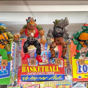 May include: Four action figures from the Teenage Mutant Ninja Turtles franchise. The figures are displayed in front of vintage basketball and football card boxes. The figures include Leonardo, Bebop, Rocksteady, and Raphael. The figures are in various colors, including green, orange, and brown.