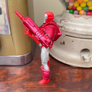 Pode incluir: Uma figura de a&ccedil;&atilde;o vermelha e prateada de um super-her&oacute;i, segurando uma arma vermelha. A figura est&aacute; em frente a uma m&aacute;quina de goma de mascar vintage e um dispensador de goma de mascar branco e transparente.