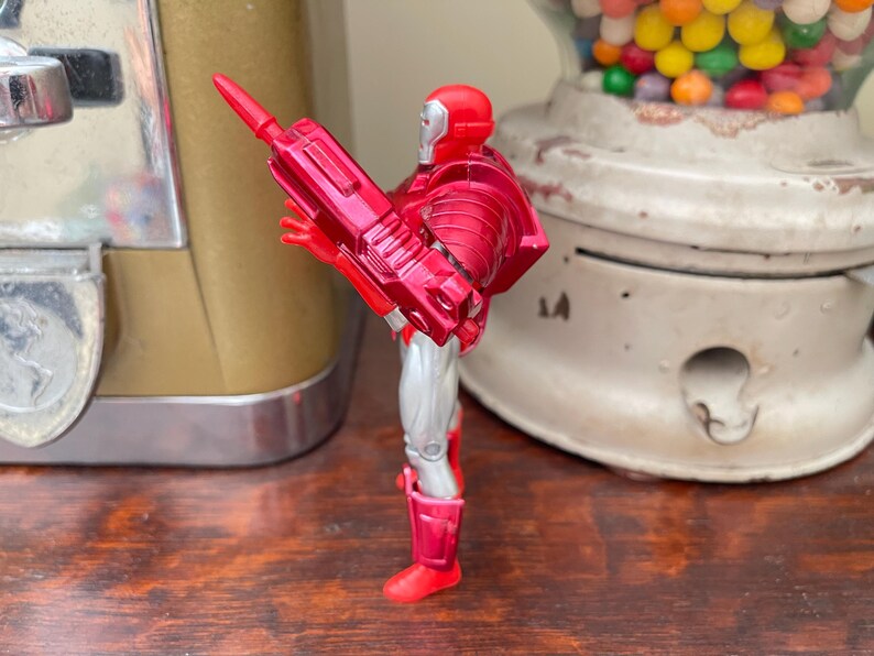 Pode incluir: Uma figura de a&ccedil;&atilde;o vermelha e prateada de um super-her&oacute;i, segurando uma arma vermelha. A figura est&aacute; em frente a uma m&aacute;quina de goma de mascar vintage e um dispensador de goma de mascar branco e transparente.