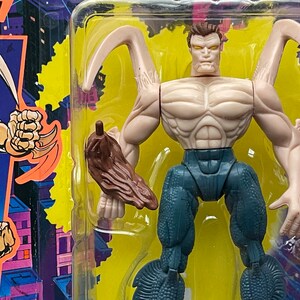 May include: A plastic action figure of a humanoid creature with a blue and gray body, brown arms, and a pale face. The figure has a pair of large, curved, brown appendages on its back. The figure is packaged in a plastic blister pack with a yellow background.