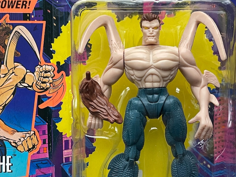 May include: A plastic action figure of a humanoid creature with a blue and gray body, brown arms, and a pale face. The figure has a pair of large, curved, brown appendages on its back. The figure is packaged in a plastic blister pack with a yellow background.