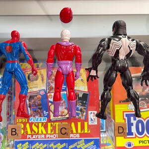 May include: Three action figures of comic book characters: Spider-Man in blue and red, Magneto in red and purple, and Venom in black and white. The figures are in front of vintage trading card boxes. Scrabble tiles spell out "BASKETBALL" and "FOOTBALL".