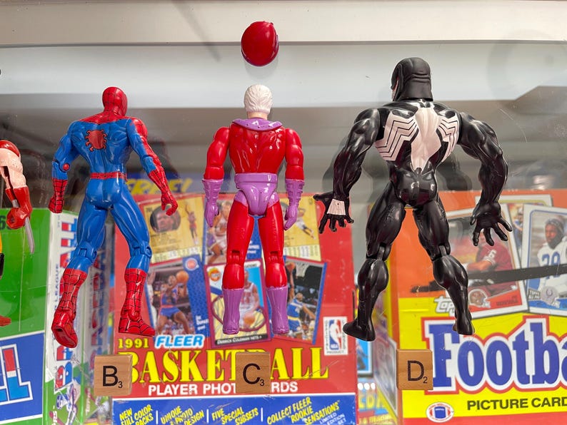 May include: Three action figures of comic book characters: Spider-Man in blue and red, Magneto in red and purple, and Venom in black and white. The figures are in front of vintage trading card boxes. Scrabble tiles spell out "BASKETBALL" and "FOOTBALL".