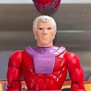 May include: A vintage action figure of a character with white hair, a stern expression, and a red and purple costume. The figure has a helmet and is from a comic book series.