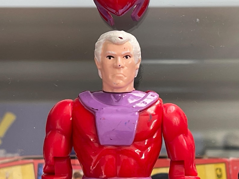 May include: A vintage action figure of a character with white hair, a stern expression, and a red and purple costume. The figure has a helmet and is from a comic book series.