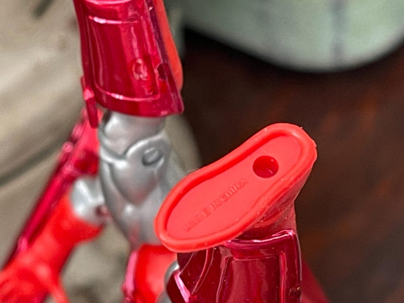 Pode incluir: Close-up de uma figura de a&ccedil;&atilde;o vermelha e prateada. O p&eacute; da figura &eacute; vermelho com um buraco e o texto "MADE IN CHINA". A perna e outras partes da figura s&atilde;o vis&iacute;veis.