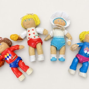 May include: Four vintage Cabbage Patch Kids dolls. The dolls have painted faces, yellow or brown hair, and wear colorful outfits. One doll wears a white bonnet and blue outfit. The other dolls wear red, blue, and white outfits.