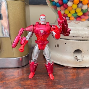Pode incluir: Uma figura de a&ccedil;&atilde;o do Homem de Ferro vermelha e prateada. A figura est&aacute; em p&eacute; com os bra&ccedil;os levantados e tem um capacete, uma placa peitoral e botas vermelhas. A figura tem aproximadamente 15 cm de altura.