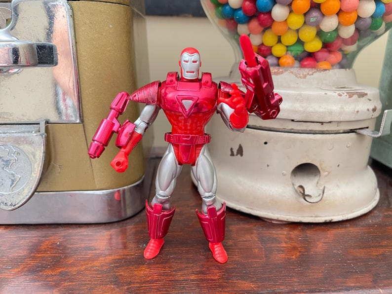 Pode incluir: Uma figura de a&ccedil;&atilde;o do Homem de Ferro vermelha e prateada. A figura est&aacute; em p&eacute; com os bra&ccedil;os levantados e tem um capacete, uma placa peitoral e botas vermelhas. A figura tem aproximadamente 15 cm de altura.