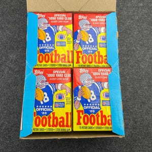 May include: A cardboard box containing four packs of Topps '89 football cards. Each pack features a football player in a blue uniform, holding a football. The packs are yellow, blue, and red.